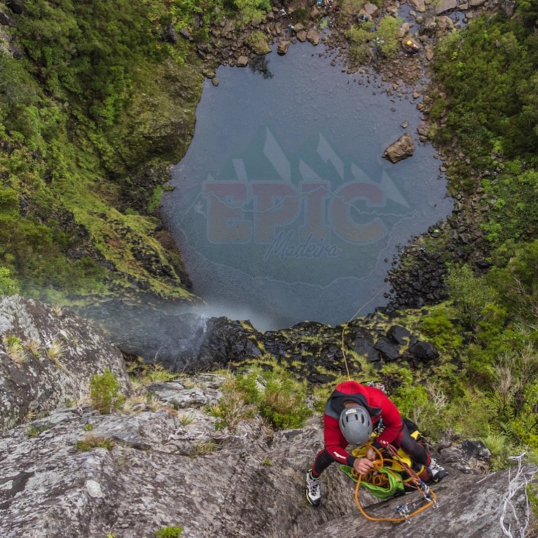 Canyoneer in a red wetsuit and helmet starting a rappel above a waterfall pool in a green Madeira canyon