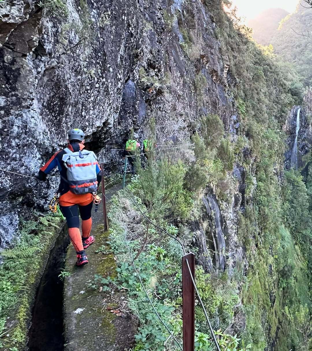 Canyoneer with helmet and backpack walking a narrow levada path along a mossy cliff, waterfall in the distance, Madeira